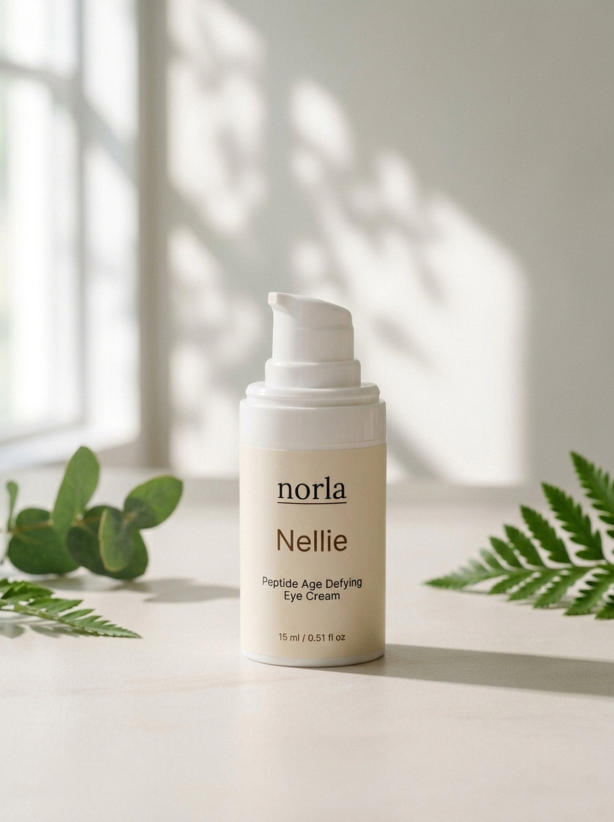 Nellie Peptide Age-Defying Eye Cream 15 ml by Norla, small airless pump bottle on a marble surface with eucalyptus and fern
