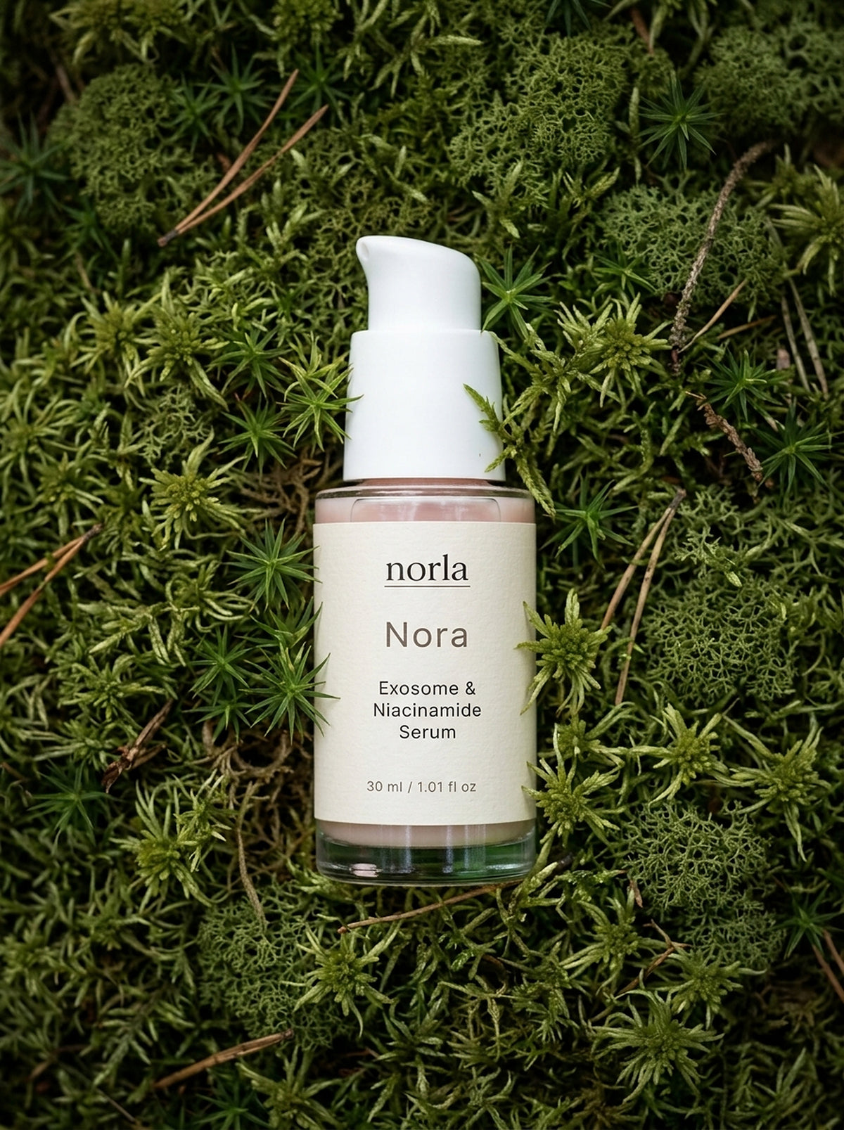 Nora Exosome & Niacinamide Serum 30 ml by Norla, blush-pink glass pump bottle lying on green moss